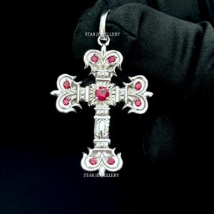 May include: A silver pendant in the shape of a cross, adorned with red gemstones. The pendant has an ornate design with intricate details and is suspended from a loop. The text "STAR JEWELLERY" is visible on the pendant.
