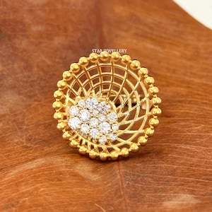 May include: A gold ring with a circular design. The ring features a lattice pattern and a cluster of small, clear gemstones at the centre. The outer edge is adorned with small, round beads. The text "STAR JEWELLERY" is visible.