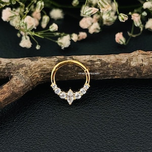 May include: Gold nose ring with a row of clear cubic zirconia stones and a larger marquise-shaped stone in the center. The ring is on a wooden surface.