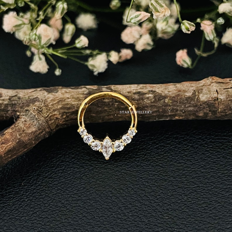 May include: Gold nose ring with a row of clear cubic zirconia stones and a larger marquise-shaped stone in the center. The ring is on a wooden surface.