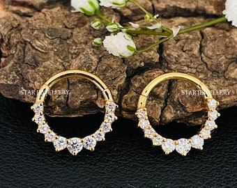May include: Two gold hoop earrings with a row of clear cubic zirconia stones around the edge. The earrings are on a wooden surface.