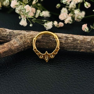 May include: Gold hoop earring with a decorative design featuring small clear stones. The hoop is on a wooden surface.