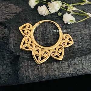 May include: Gold hoop earring with a delicate, intricate design featuring a series of connected swirls and hearts. The earring is on a dark wooden surface.