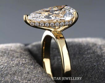 Könnte beinhalten: Ein Ring aus Gelbgold mit einem großen birnenförmigen Mittelstein und einem versteckten Heiligenschein aus kleinen runden Steinen, die entlang des Korbes gefasst sind. Die Ringschiene ist glatt und poliert. Der Text STAR JEWELLERY ist sichtbar.