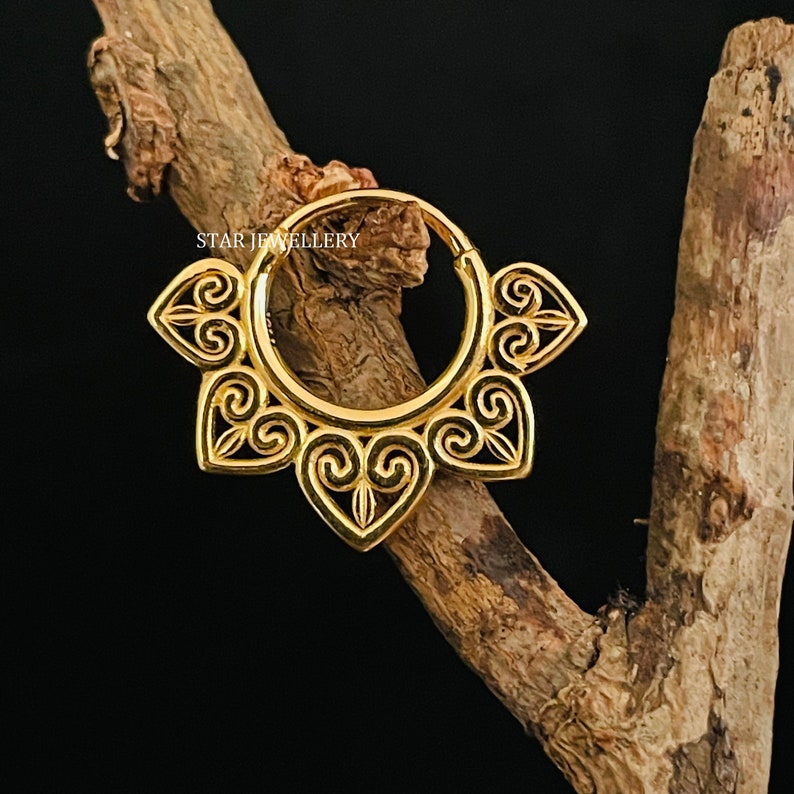 May include: Gold-toned hoop earring with a decorative design featuring intricate swirls and heart shapes. The earring is made of metal and has a circular shape.