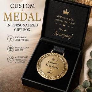 Puede incluir: Una medalla personalizada dorada en una caja de regalo negra. La medalla tiene un diseño de corona de laurel y espacio para texto personalizado. La caja abierta revela la medalla y las palabras "To the one who inspires every day, You are Amazing!"