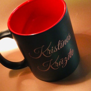 May include: Black coffee mug with a red interior and a black handle. The mug has the name "Kristines Krūzīte" in a cursive font on the side. The mug is angled slightly, showing the red interior.
