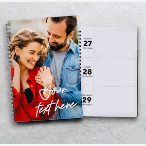 May include: A spiral-bound planner with a photograph of a smiling couple on the cover. The cover has the text "Your text here" in a handwritten style. The open planner shows the dates December 27, 28, and 29.