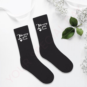Può includere: Un paio di calzini neri con la scritta bianca "2 Amazing years" e la data "27.07.2017". I calzini sono presentati su uno sfondo bianco con decorazioni floreali.