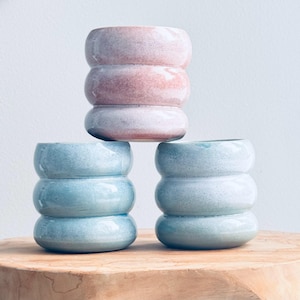 Könnte beinhalten: Drei Keramikvasen in einem gestapelten, abgerundeten Design. Eine Vase ist rosa und zwei sind blau, alle mit einer gesprenkelten Glasur. Die Vasen stehen auf einer Holzoberfläche vor einem weißen Hintergrund.