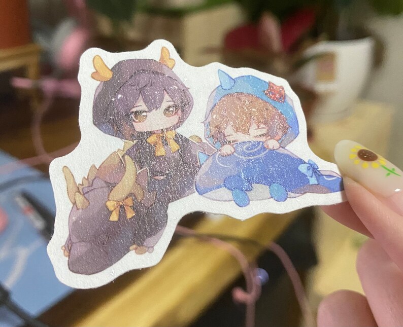 Puede incluir: Dos personajes de dibujos animados, uno con un disfraz con capucha azul y blanco y el otro con un disfraz negro y amarillo, est&aacute;n representados en una pegatina. La pegatina es blanca con una superficie ligeramente texturizada.