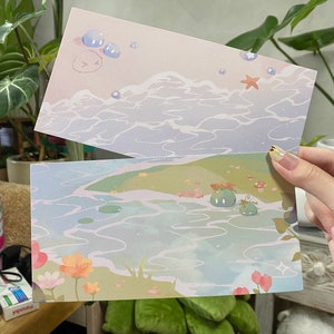 Pode incluir: Dois postais ilustrados. O cartão superior retrata uma cena oceânica pastel com bolhas e uma estrela do mar. O cartão inferior mostra uma paisagem serena com água, flores e criaturas de desenho animado. Os cartões são segurados.