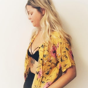 May include: Yellow floral button-down shirt with pink and green accents. The shirt is paired with a black bra and black bottoms. The image includes the text "plat_arte" in the bottom left corner.