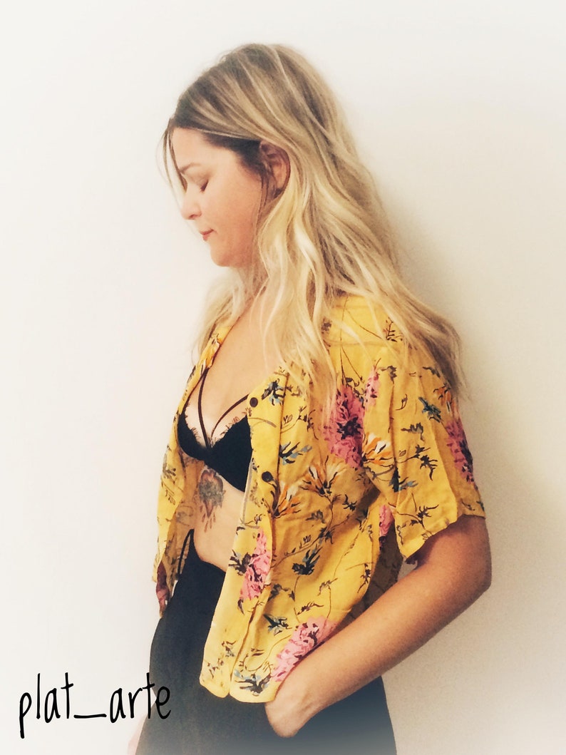 May include: Yellow floral button-down shirt with pink and green accents. The shirt is paired with a black bra and black bottoms. The image includes the text "plat_arte" in the bottom left corner.