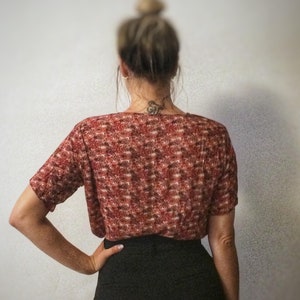 May include: A short-sleeved, red and brown patterned top paired with a black skirt. The top has a textured appearance. The image is taken from behind, showcasing the back of the outfit. The brand name "plat_arte" is visible in the bottom left corner.