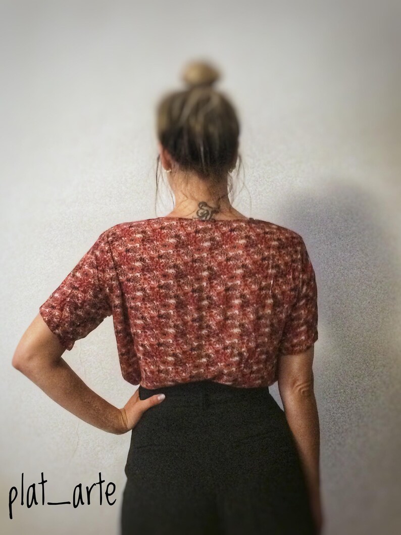 May include: A short-sleeved, red and brown patterned top paired with a black skirt. The top has a textured appearance. The image is taken from behind, showcasing the back of the outfit. The brand name "plat_arte" is visible in the bottom left corner.