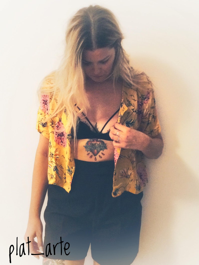 May include: A yellow floral print short-sleeved shirt with pink and green accents. The shirt is paired with black shorts and a black bralette. The image has a vintage aesthetic, with the text "plat_arte" in the bottom left corner.