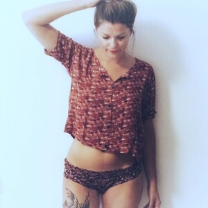 May include: A cropped, button-down shirt in a reddish-brown pattern paired with dark patterned underwear. A tattoo of a bird is visible on the thigh. The image is set against a plain white background.