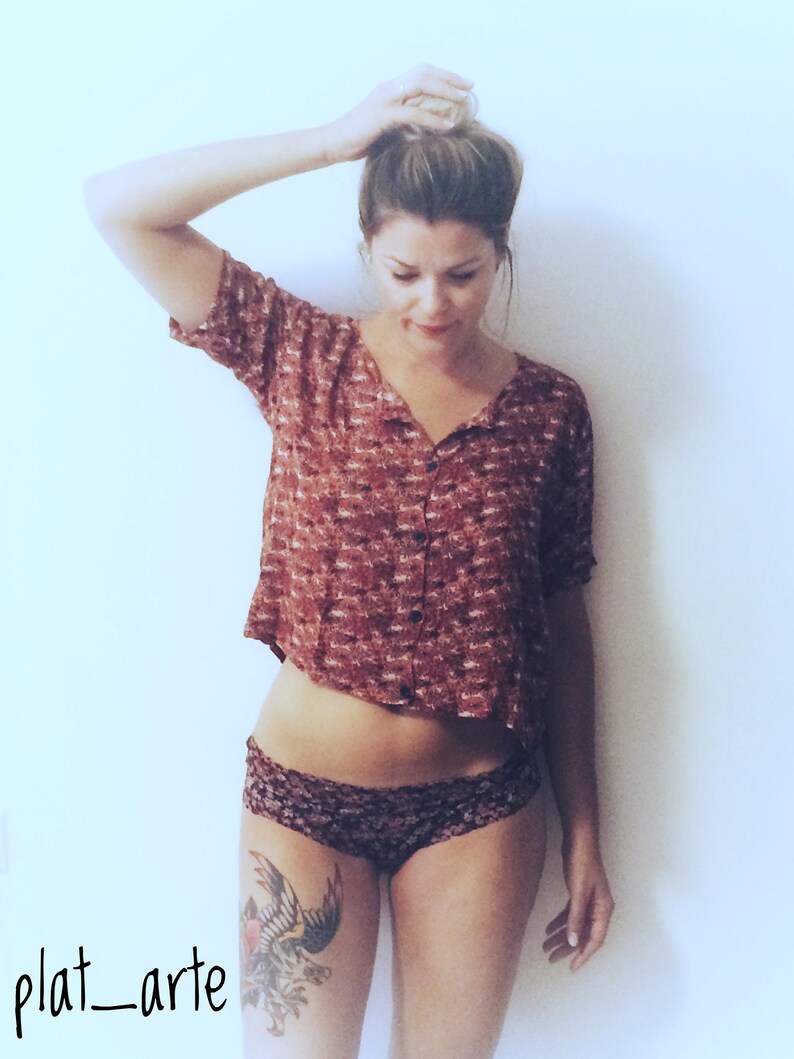 May include: A cropped, button-down shirt in a reddish-brown pattern paired with dark patterned underwear. A tattoo of a bird is visible on the thigh. The image is set against a plain white background.