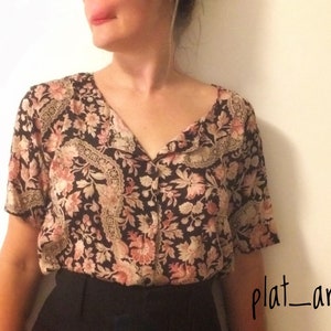 May include: A short-sleeved blouse with a floral pattern in shades of black, pink, and beige. The blouse has a V-neckline and decorative trim along the collar and front. The bottom half of the image shows black pants.