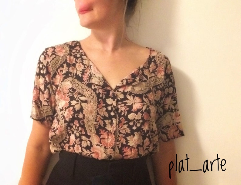 May include: A short-sleeved blouse with a floral pattern in shades of black, pink, and beige. The blouse has a V-neckline and decorative trim along the collar and front. The bottom half of the image shows black pants.