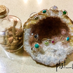 May include: A collection of gemstone rings displayed in a geode. The rings are made of silver and feature various gemstones, including turquoise, green, red, and black. The geode is a natural rock formation with a pink and white interior.
