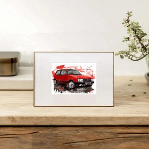 May include: A rectangular white picture frame containing a watercolor illustration of a red hatchback car. The artwork features expressive red and black paint splatters surrounding the vehicle, which is depicted from a front three-quarter angle.