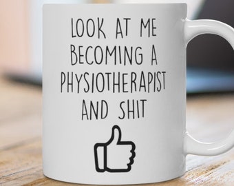 May include: White ceramic mug featuring the text "LOOK AT ME BECOMING A PHYSIOTHERAPIST AND SHIT" in black, and a thumbs-up graphic. The mug sits on a wooden surface, with a laptop visible in the background.