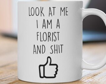 May include: White ceramic mug with the text "LOOK AT ME I AM A FLORIST AND SHIT" in black, along with a thumbs-up graphic. The mug is on a wooden surface, with a laptop in the background.