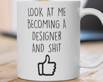 May include: White ceramic mug with the text "LOOK AT ME BECOMING A DESIGNER AND SHIT" in black, along with a thumbs-up graphic. The mug is on a wooden surface, with a laptop in the background.