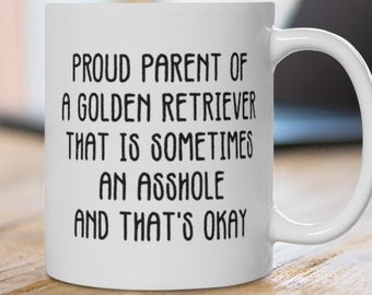 May include: White ceramic mug with the text "PROUD PARENT OF A GOLDEN RETRIEVER THAT IS SOMETIMES AN ASSHOLE AND THAT'S OKAY" in black. The mug is on a wooden table with a laptop in the background.