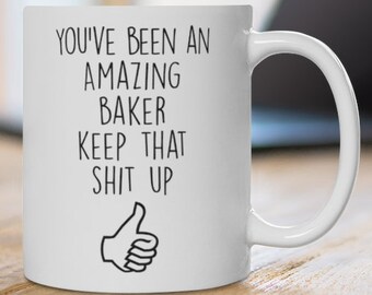 May include: White ceramic mug with the text "YOU'VE BEEN AN AMAZING BAKER KEEP THAT SHIT UP" in black, along with a thumbs-up graphic. The mug is on a wooden surface, with a laptop in the background.