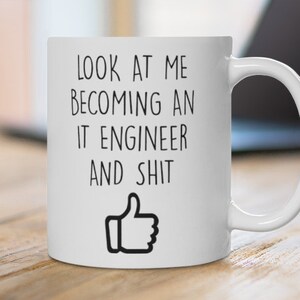 Könnte beinhalten: Weiße Keramik-Tasse mit dem Text "LOOK AT ME BECOMING AN IT ENGINEER AND SHIT" in Schwarz und einem Daumen-hoch-Symbol. Die Tasse steht auf einer Holzoberfläche, mit einem Laptop im Hintergrund.