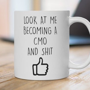 May include: White ceramic coffee mug with the text "LOOK AT ME BECOMING A CMO AND SHIT" in black, along with a thumbs-up graphic. The mug is on a wooden surface, with a laptop in the background.