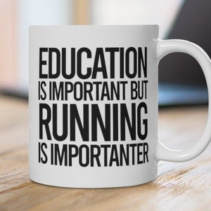 May include: White ceramic mug with the text "EDUCATION IS IMPORTANT BUT RUNNING IS IMPORTER". The mug is on a wooden surface with a laptop in the background. The mug has a curved handle.