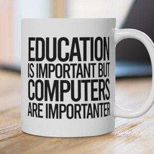 Puede incluir: Taza de cerámica blanca con el texto "EDUCATION IS IMPORTANT BUT COMPUTERS ARE IMPORTER". La taza tiene un asa curva y está sobre una superficie de madera. Un portátil está en el fondo.