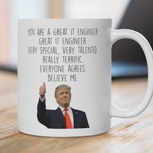 May include: White ceramic mug with a handle. The mug has the text "YOU ARE A GREAT IT ENGINEER. GREAT IT ENGINEER. VERY SPECIAL, VERY TALENTED. REALLY TERRIFIC. EVERYONE AGREES. BELIEVE ME." and an image of a man giving a thumbs up.