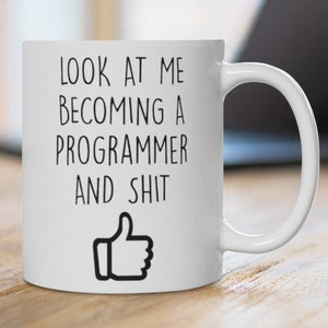 May include: White ceramic mug with the text "LOOK AT ME BECOMING A PROGRAMMER AND SHIT" in black, and a thumbs-up graphic. The mug is on a wooden surface, with a laptop in the background.