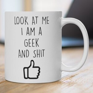 May include: White ceramic mug with the text "LOOK AT ME I AM A GEEK AND SHIT" in black, and a thumbs-up graphic. The mug is on a wooden surface, with a laptop in the background.