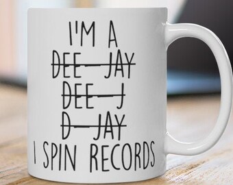 May include: White ceramic mug with the text "I'M A DEE-JAY DEE-J D-JAY I SPIN RECORDS" in black. The mug is on a wooden surface with a laptop in the background. The mug has a curved handle.