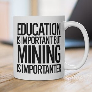 Könnte beinhalten: Weiße Keramik-Tasse mit dem Text "EDUCATION IS IMPORTANT BUT MINING IS IMPORTER". Die Tasse steht auf einer Holzoberfläche, im Hintergrund ein Laptop. Die Tasse hat einen weißen Henkel.