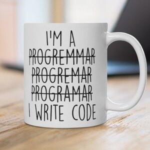 May include: White ceramic mug with the text "I'M A PROGRAMMAR PROGRAMMAR PROGRAMMAR I WRITE CODE" in black. The mug is on a wooden surface, with a laptop in the background.