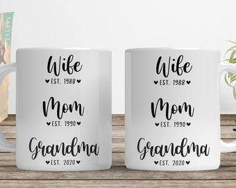 May include: Two white ceramic mugs with black text that reads "Wife Est. 1988, Mom Est. 1990, Grandma Est. 2020". The mugs are on a wooden surface, with books and a small plant in the background.