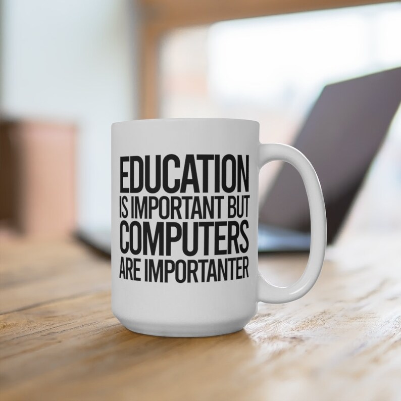 May include: White ceramic mug with a large handle. The mug features the text "EDUCATION IS IMPORTANT BUT COMPUTERS ARE IMPORTER". A laptop is visible in the background. The mug is on a wooden surface.