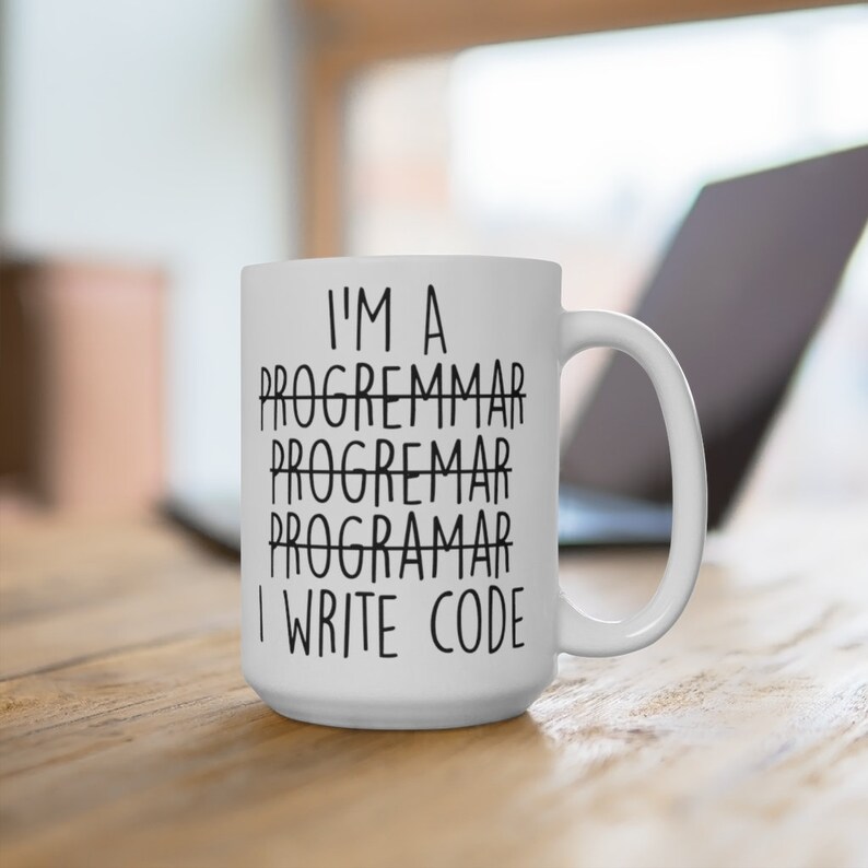 May include: White ceramic mug with the text "I'M A PROGRAMMAR PROGRAMMAR PROGRAMAR I WRITE CODE" in black. The mug is on a wooden surface with a laptop in the background. The mug has a large handle.