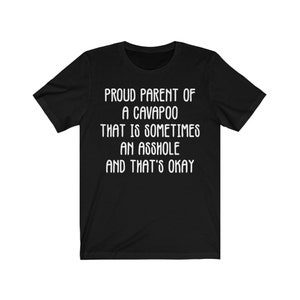 May include: Black t-shirt with white text that reads: "PROUD PARENT OF A CAVAPOO THAT IS SOMETIMES AN ASSHOLE AND THAT'S OKAY." The shirt has short sleeves and a crew neck.