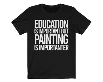May include: Black t-shirt with white text. The text reads: "EDUCATION IS IMPORTANT BUT PAINTING IS IMPORTER." The shirt is a crew neck style and appears to be made of a soft material. The shirt is a casual wear item.