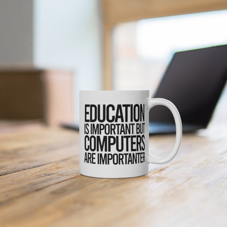 May include: White ceramic mug with the text "EDUCATION IS IMPORTANT BUT COMPUTERS ARE IMPORTER". The mug has a curved handle and is sitting on a wooden surface. A laptop is in the background.