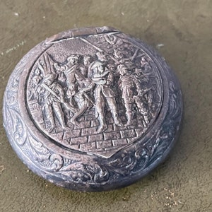 Puede incluir: Un compacto plateado con un diseño ornamentado que presenta una imagen en relieve de un grupo de personas en procesión. El compacto tiene una tapa con bisagras y un borde floral detallado.