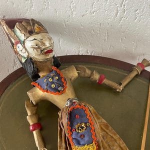 Peut inclure: Une marionnette à tige en bois avec un visage peint à la main, des cheveux noirs, une bouche rouge et une peau blanche. La figurine porte un plastron et une jupe en tissu bleu avec des bordures orange, des paillettes dorées et des franges jaunes.